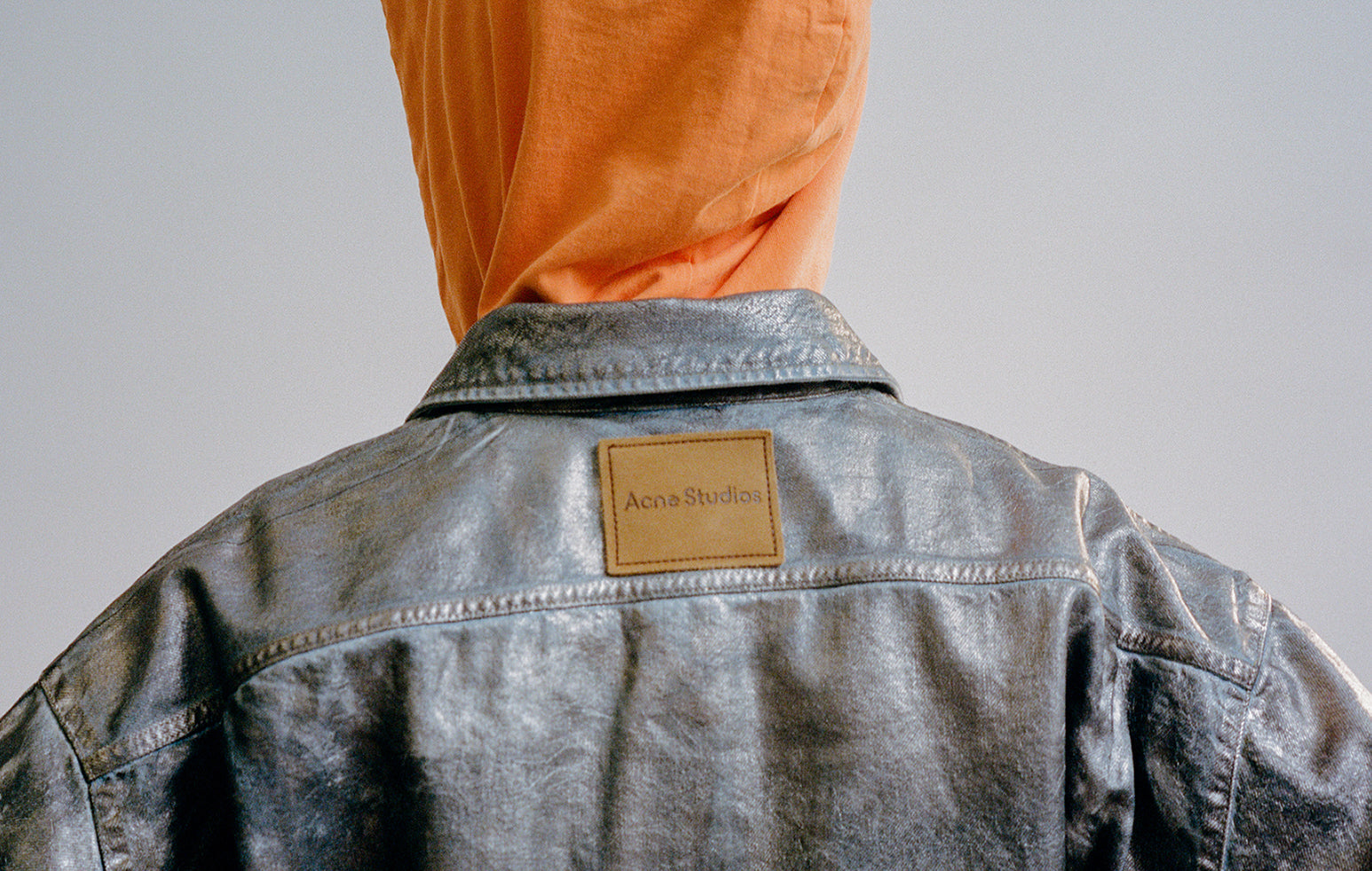 A man standing with his back turned, wearing a hood and a denim jacket